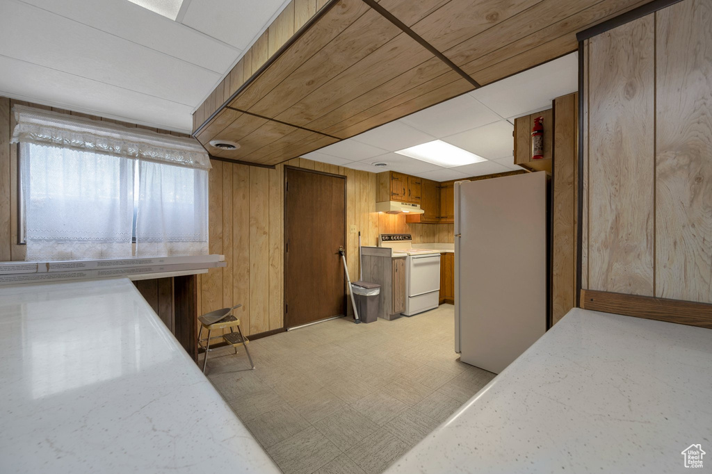 Kitchen featuring white appliances, under cabinet range hood, light flooring, wooden walls, and brown cabinetry