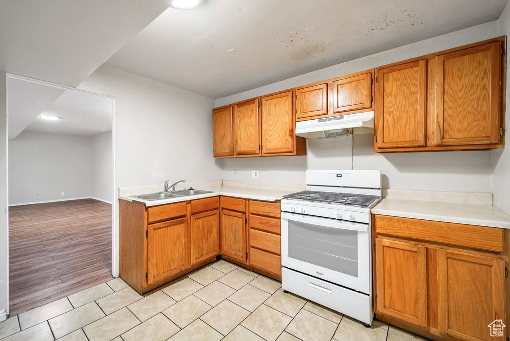 Kitchen featuring white range with gas stovetop, under cabinet range hood, a sink, light countertops, and brown cabinets