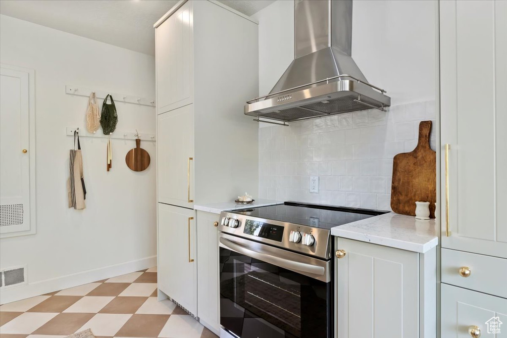 Kitchen with wall chimney exhaust hood, stainless steel electric range, light floors, decorative backsplash, and baseboards