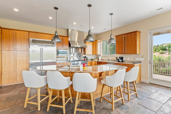 Kitchen featuring wall chimney range hood, built in appliances, stone tile floors, a sink, and recessed lighting