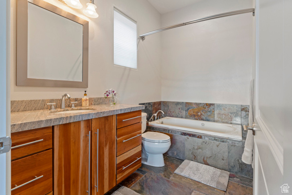 Full bath with stone finish flooring, vanity, a relaxing tiled tub, and toilet