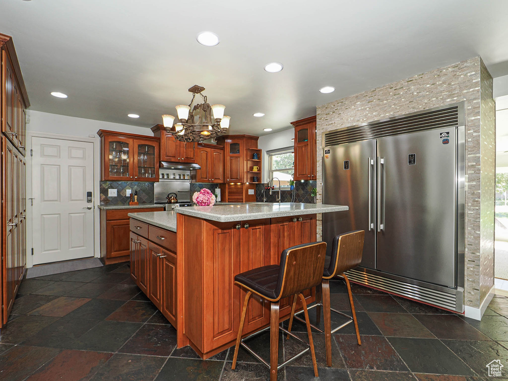 Kitchen with built in refrigerator, a chandelier, under cabinet range hood, decorative backsplash, and a kitchen island