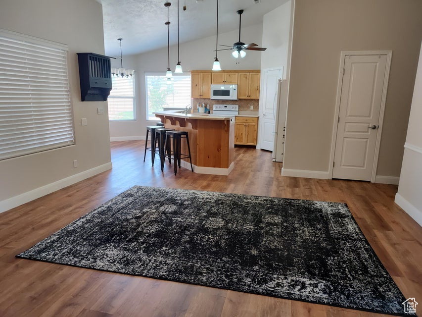 Kitchen with white microwave, light brown cabinetry, vaulted ceiling, an island with sink, and baseboards