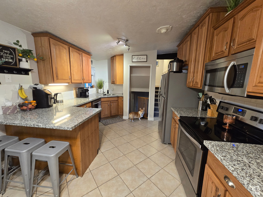 Kitchen with stainless steel appliances, brown cabinetry, light stone countertops, a peninsula, and a textured ceiling