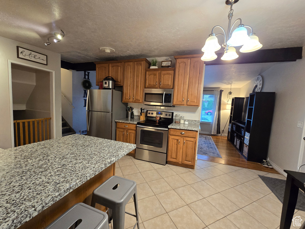 Kitchen with appliances with stainless steel finishes, light tile patterned flooring, a chandelier, brown cabinets, and light stone counters