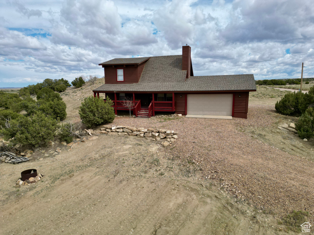 View of front of home featuring covered porch, dirt driveway, an attached garage, a shingled roof, and a chimney