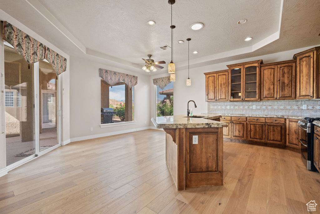 Kitchen featuring a tray ceiling, a sink, double oven range, brown cabinetry, and a center island with sink