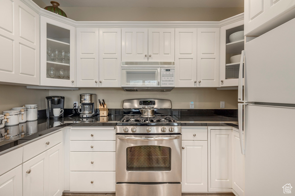 Kitchen with white appliances, white cabinets, dark countertops, and glass insert cabinets