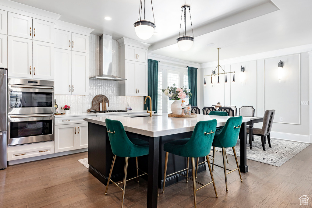 Kitchen featuring wall chimney exhaust hood, double oven, a sink, tasteful backsplash, and a breakfast bar area