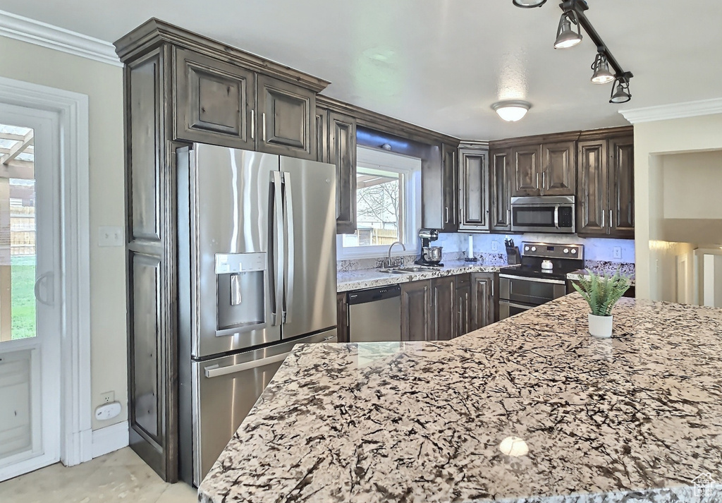 Kitchen with appliances with stainless steel finishes, a sink, light stone counters, ornamental molding, and dark brown cabinetry
