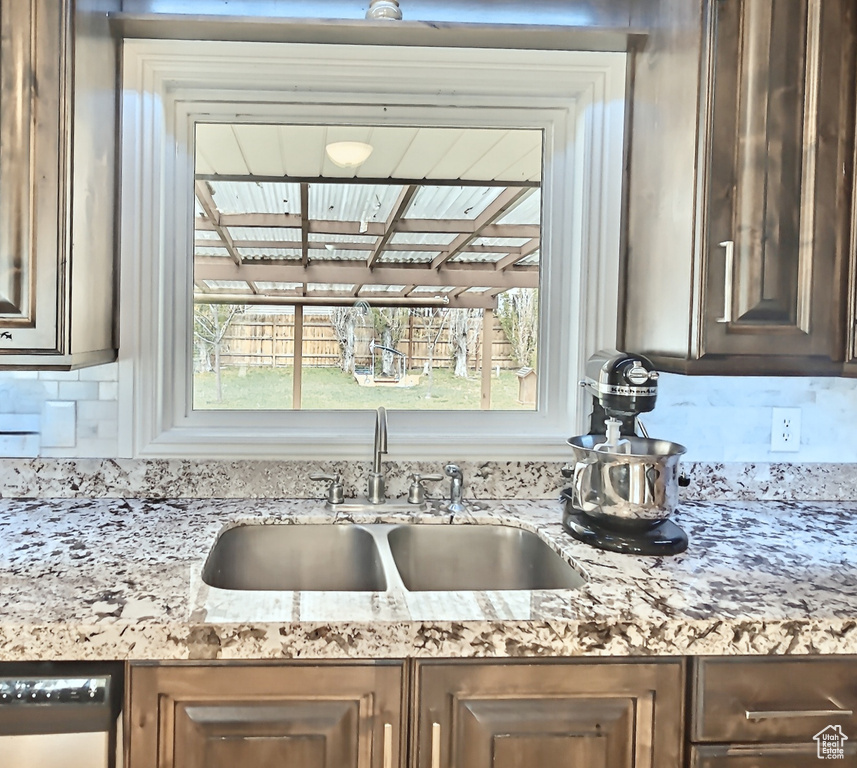 Kitchen view of a sink, decorative backsplash, dark brown cabinets, dishwasher, and light stone counters