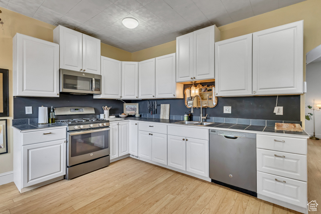 Kitchen featuring stainless steel appliances, a sink, light wood finished floors, white cabinetry, and tile counters