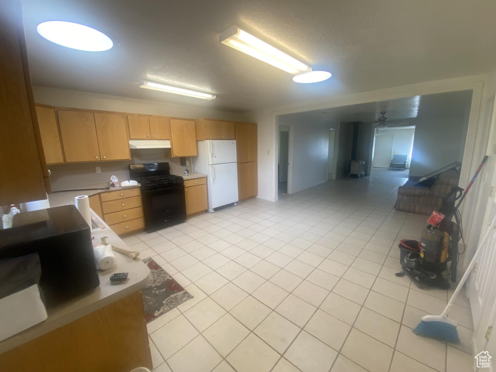 Kitchen with freestanding refrigerator, black gas range, under cabinet range hood, light brown cabinets, and light tile patterned flooring