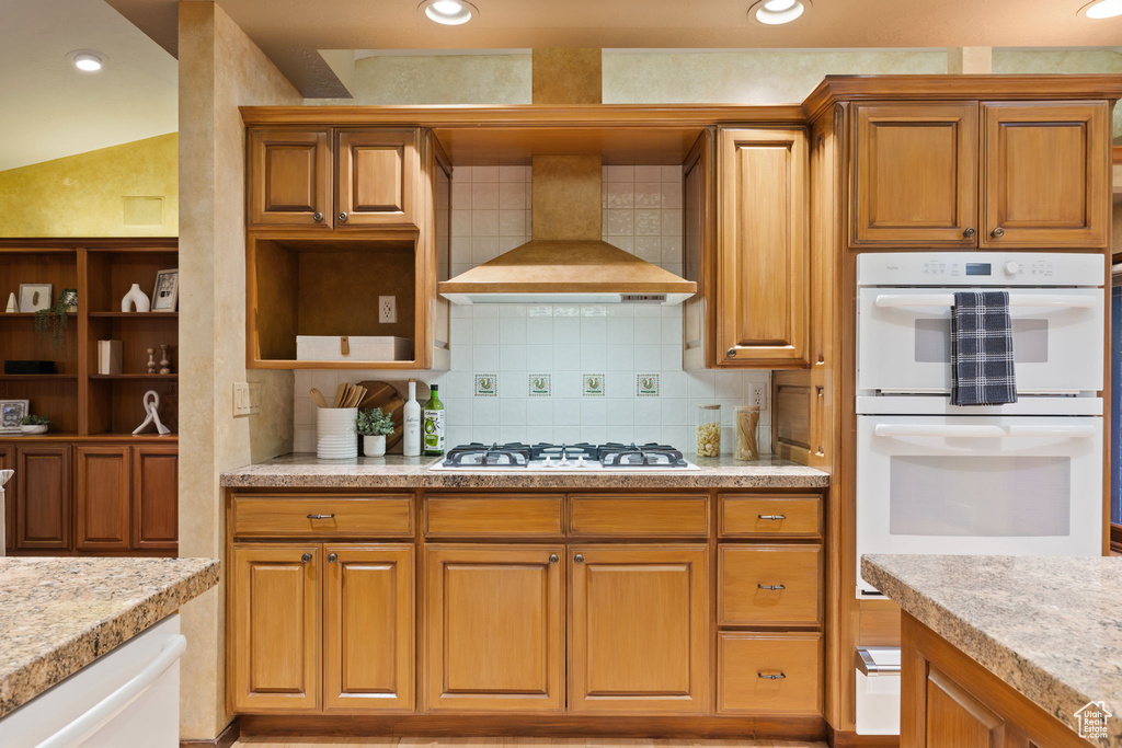 Kitchen featuring recessed lighting, brown cabinetry, white appliances, wall chimney exhaust hood, and tasteful backsplash