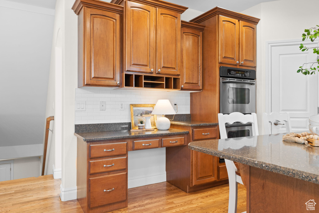 Kitchen featuring oven, backsplash, dark stone counters, light wood-style flooring, and brown cabinetry
