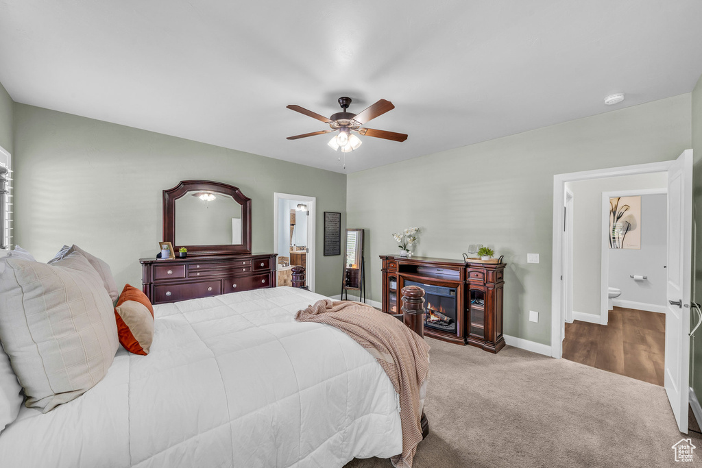 Bedroom with connected bathroom, baseboards, carpet, a ceiling fan, and a warm lit fireplace