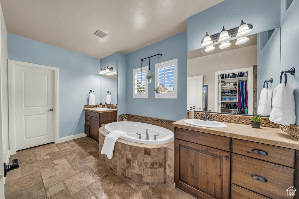 Bathroom with a garden tub, two vanities, stone tile floors, a walk in closet, and baseboards