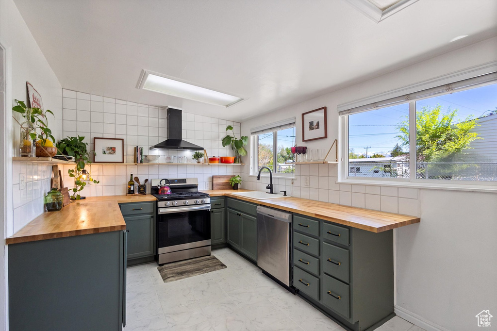 Kitchen featuring a sink, wall chimney range hood, butcher block counters, open shelves, and appliances with stainless steel finishes
