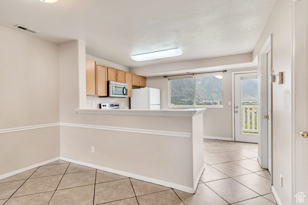 Kitchen featuring stainless steel microwave, freestanding refrigerator, light tile patterned flooring, light brown cabinets, and healthy amount of natural light