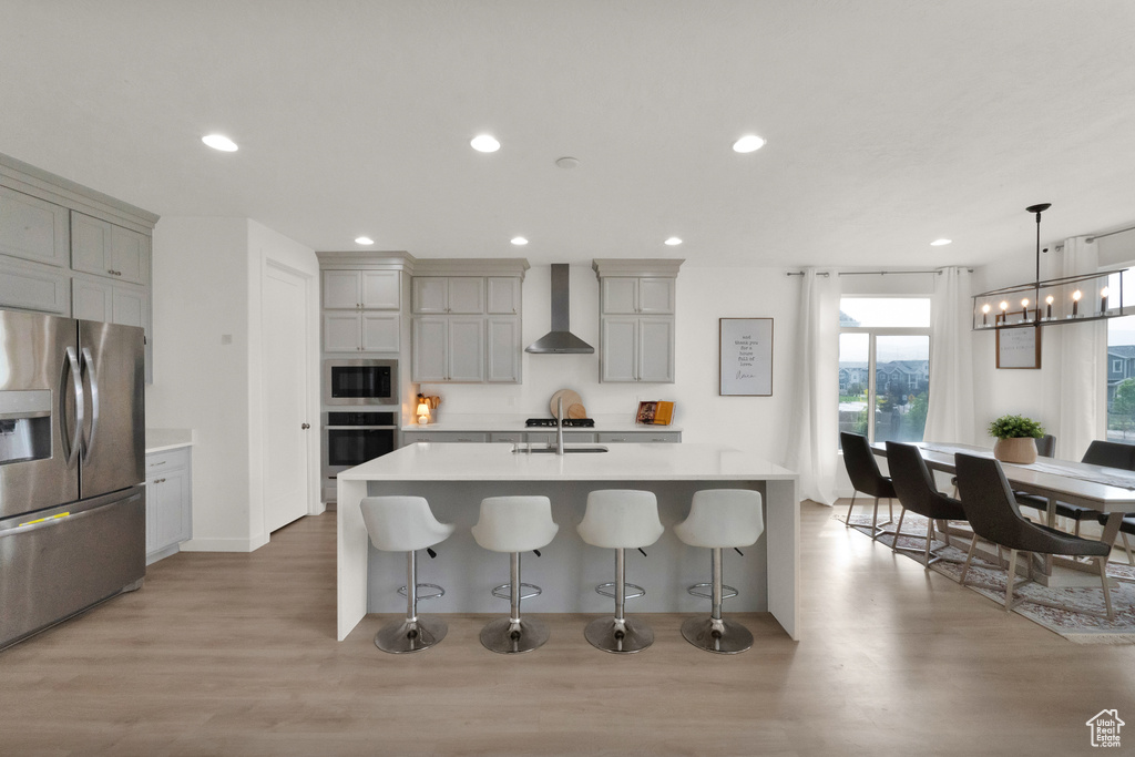 Kitchen featuring stainless steel appliances, wall chimney exhaust hood, light wood-style floors, light countertops, and a breakfast bar area