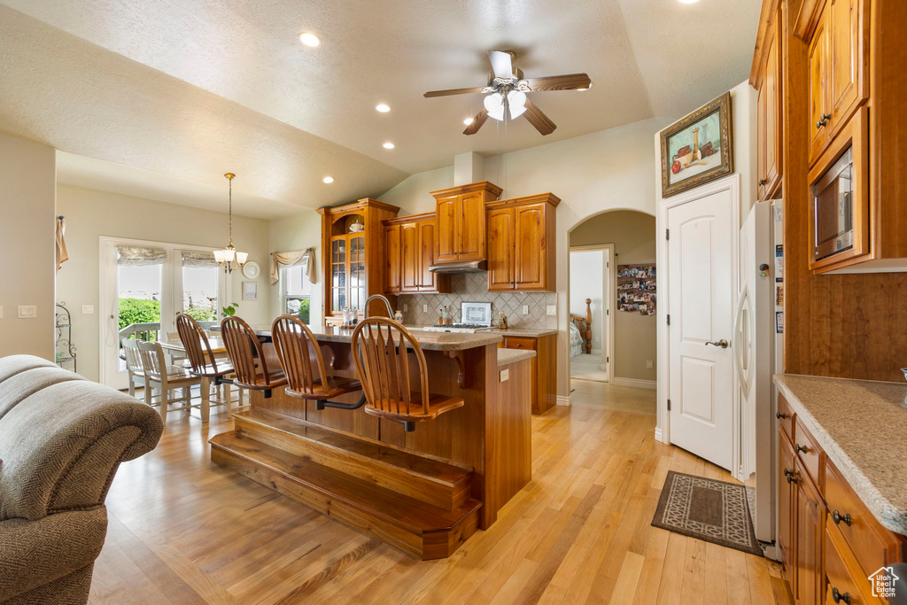 Kitchen featuring a breakfast bar, arched walkways, brown cabinetry, ceiling fan with notable chandelier, and tasteful backsplash