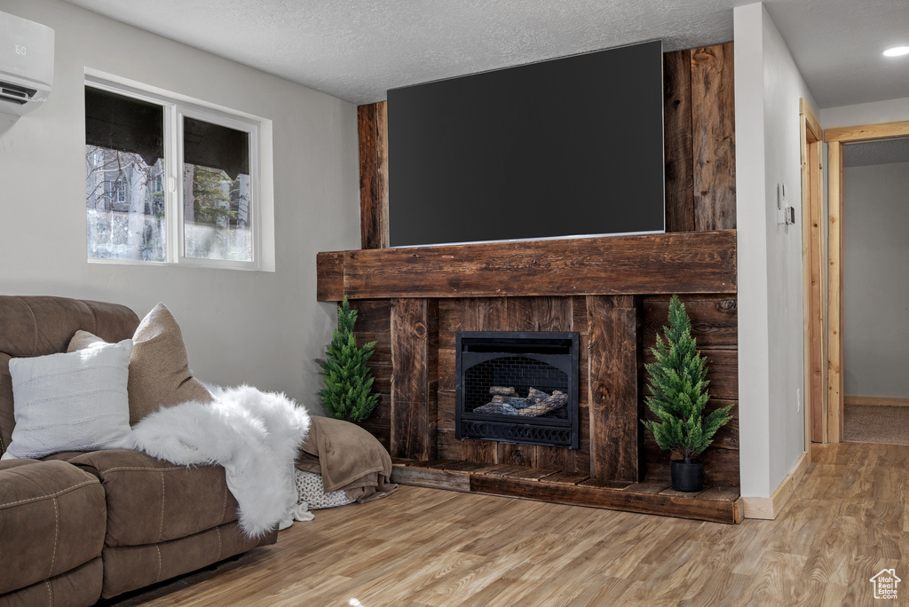 Living room with a fireplace with raised hearth, wood finished floors, a wall unit AC, a textured ceiling, and baseboards