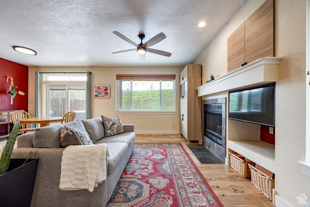 Living room featuring a textured ceiling, light wood-style flooring, a tile fireplace, ceiling fan, and baseboards