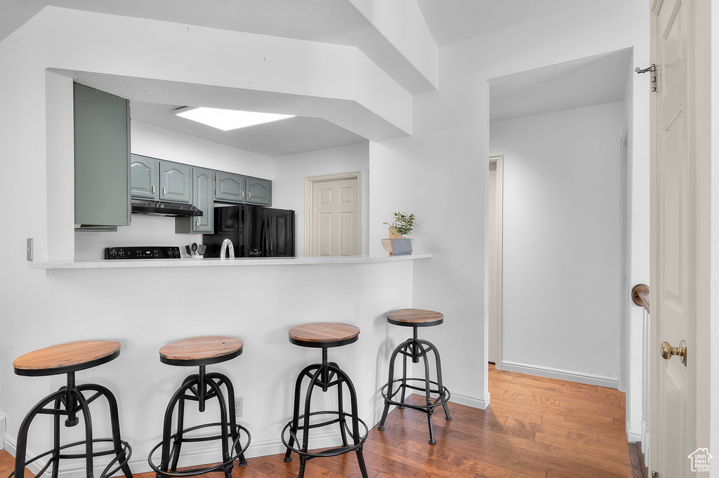 Kitchen featuring black fridge, under cabinet range hood, a breakfast bar, dark wood-type flooring, and gray cabinetry