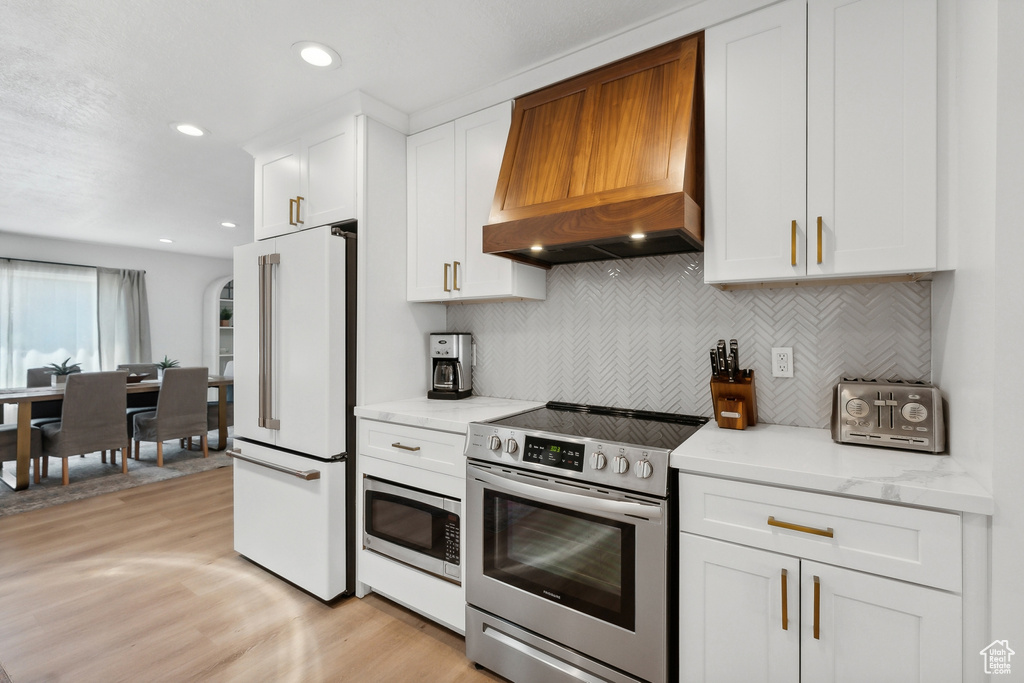 Kitchen featuring appliances with stainless steel finishes, premium range hood, white cabinetry, light wood-style flooring, and backsplash