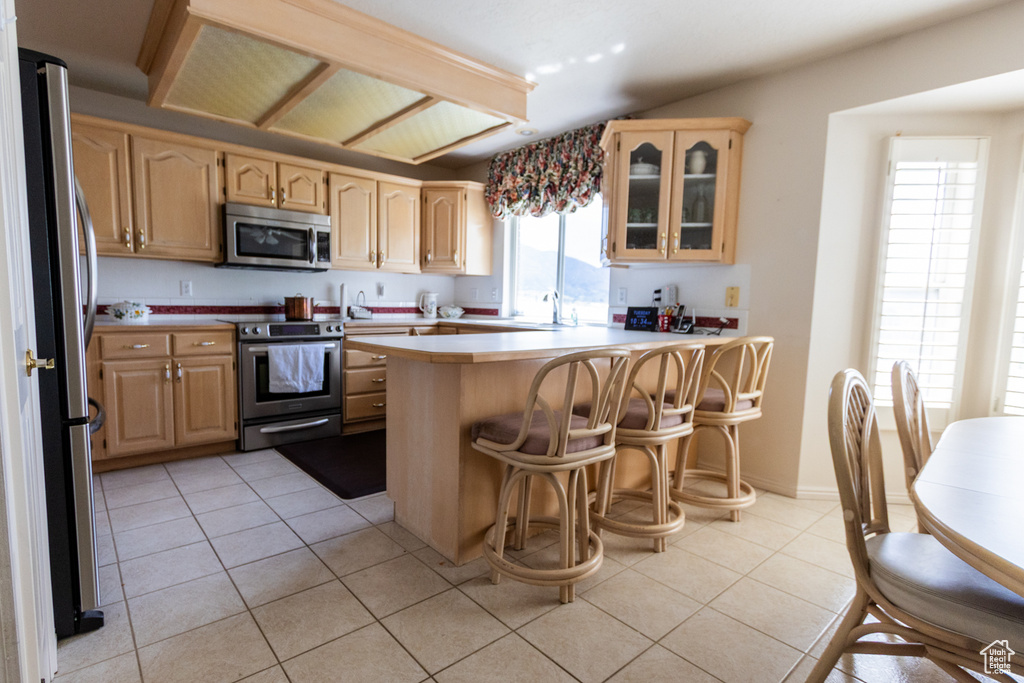 Kitchen featuring appliances with stainless steel finishes, a peninsula, light countertops, light brown cabinets, and glass insert cabinets
