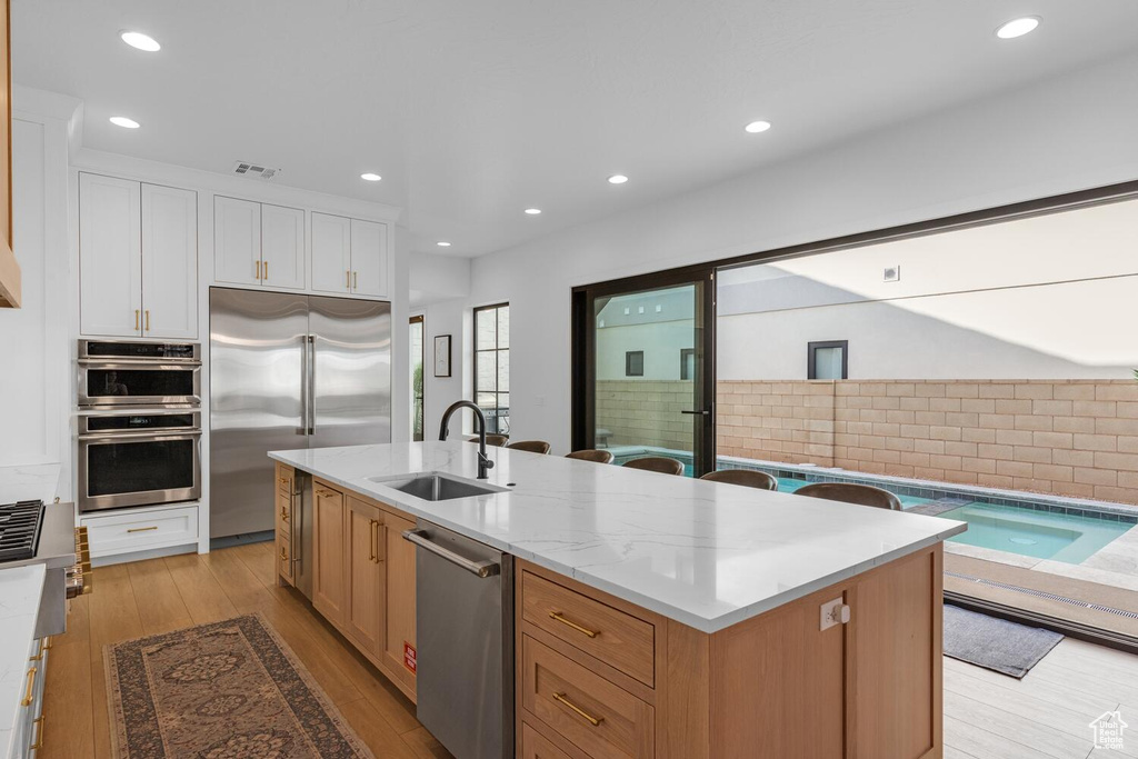 Kitchen featuring a sink, stainless steel appliances, white cabinets, a center island with sink, and light wood-style floors