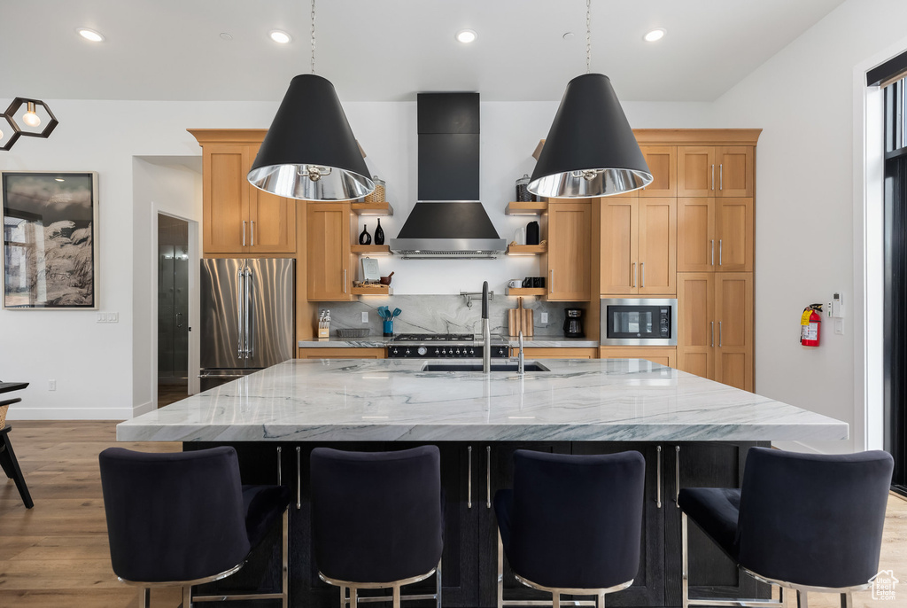 Kitchen with wall chimney exhaust hood, freestanding refrigerator, built in microwave, light wood finished floors, and a kitchen island with sink