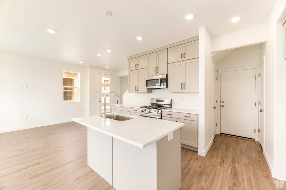 Kitchen featuring stainless steel appliances, a sink, a center island with sink, light wood-style floors, and light countertops