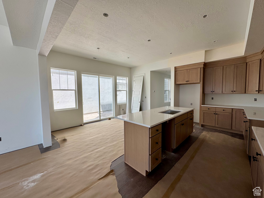 Kitchen with a textured ceiling, a center island with sink, light countertops, and brown cabinetry