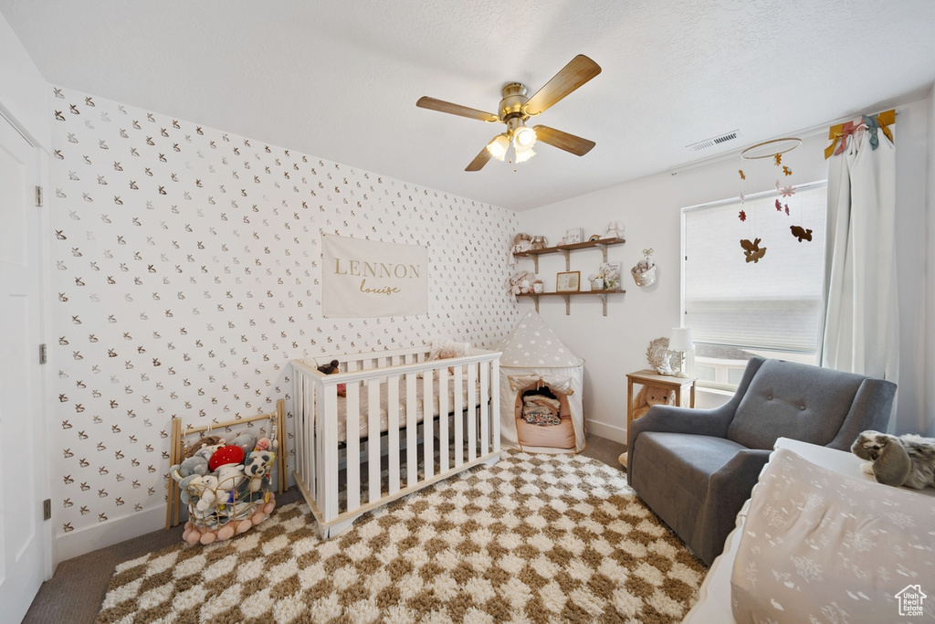 Carpeted bedroom featuring wallpapered walls, baseboards, a nursery area, ceiling fan, and an accent wall