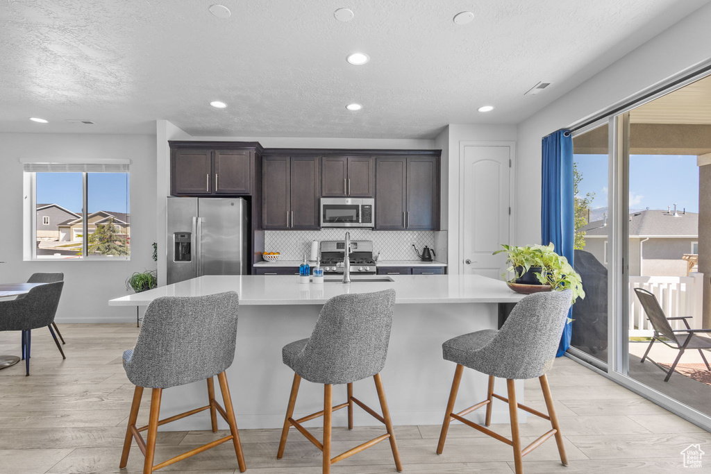 Kitchen featuring stainless steel appliances, a sink, a textured ceiling, dark brown cabinetry, and an island with sink