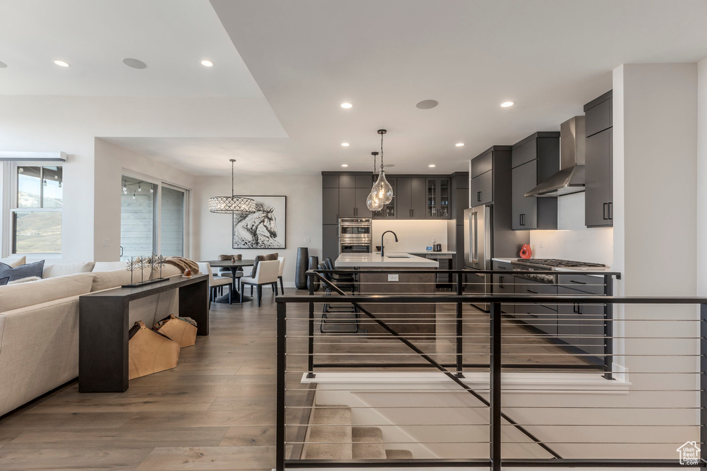 Kitchen with wall chimney exhaust hood, wood finished floors, recessed lighting, a center island with sink, and hanging light fixtures
