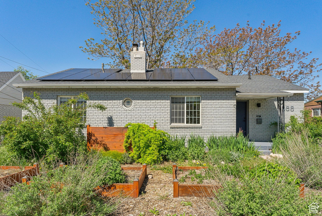 Rear view of property featuring a garden, a chimney, brick siding, a shingled roof, and solar panels