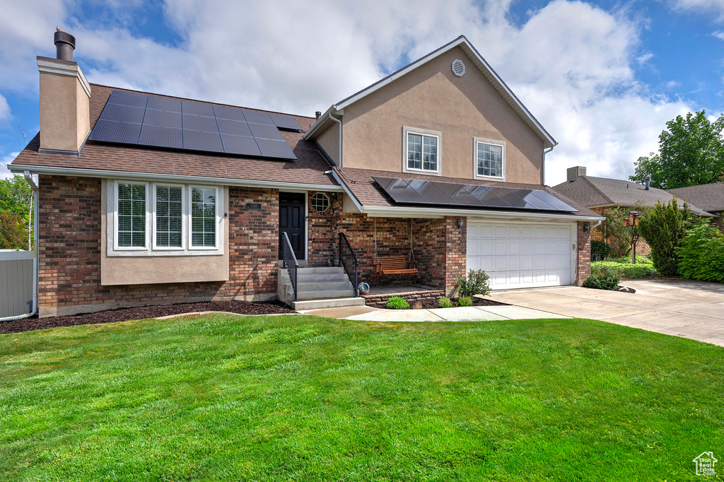 Split level home with a chimney, driveway, a garage, a front yard, and roof mounted solar panels