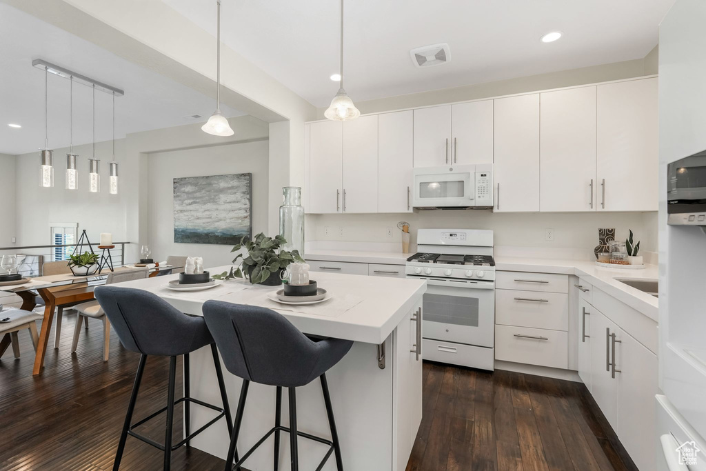 Kitchen with white appliances, dark wood-type flooring, a breakfast bar area, hanging light fixtures, and recessed lighting