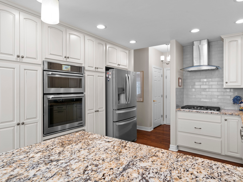 Kitchen featuring wall chimney exhaust hood, appliances with stainless steel finishes, dark wood-style flooring, backsplash, and light stone countertops