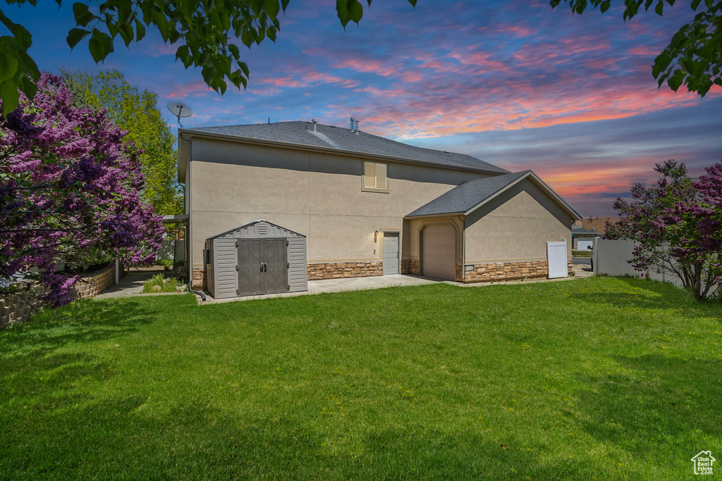 Back of house at dusk with stucco siding, a storage shed, an attached garage, stone siding, and an outdoor structure
