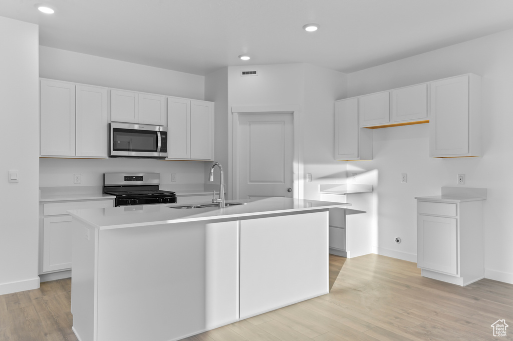 Kitchen with appliances with stainless steel finishes, a sink, white cabinetry, an island with sink, and light wood-style flooring