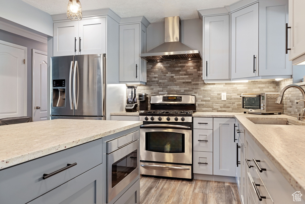 Kitchen featuring wall chimney range hood, stainless steel appliances, decorative backsplash, a sink, and light stone counters