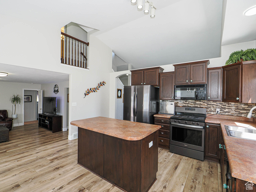 Kitchen with appliances with stainless steel finishes, a sink, light wood finished floors, dark brown cabinetry, and high vaulted ceiling
