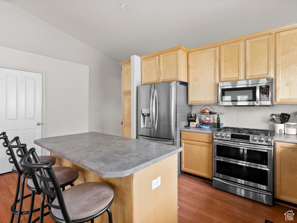 Kitchen with appliances with stainless steel finishes, light brown cabinetry, lofted ceiling, wood finished floors, and a kitchen breakfast bar