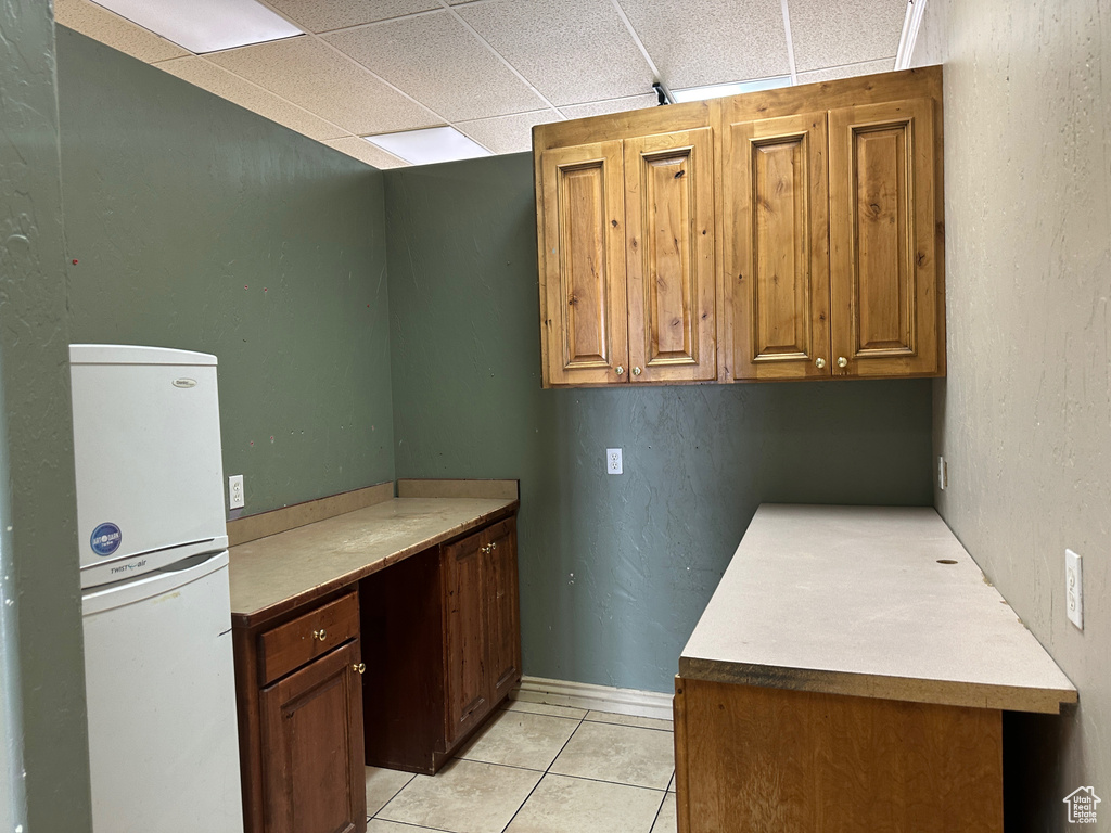 Kitchen featuring freestanding refrigerator, light tile patterned floors, a drop ceiling, light countertops, and brown cabinetry