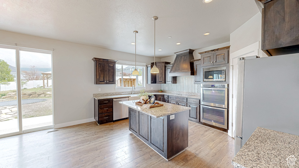 Kitchen with appliances with stainless steel finishes, custom exhaust hood, dark brown cabinetry, light wood-style flooring, and decorative light fixtures