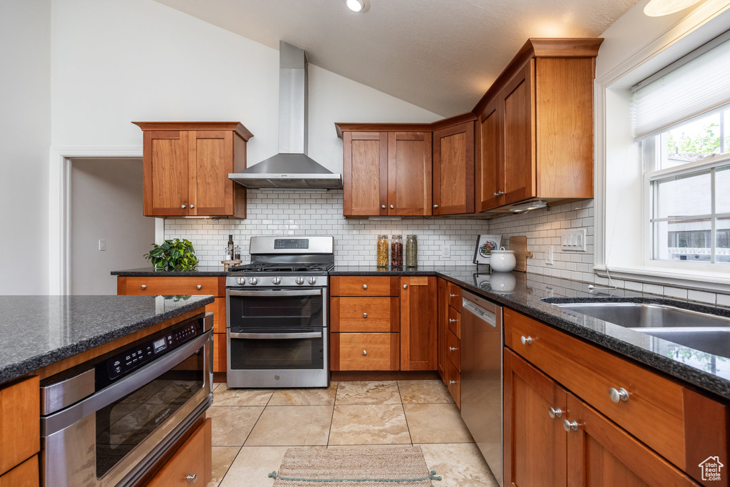 Kitchen with wall chimney range hood, stainless steel appliances, lofted ceiling, backsplash, and brown cabinetry