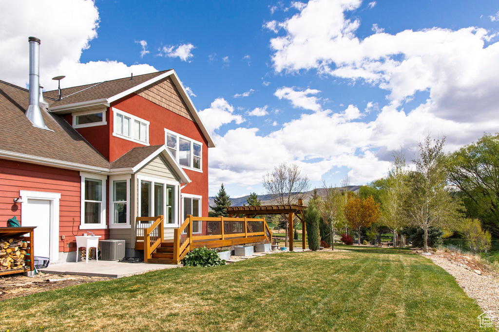 Back of property with cooling unit, a deck, a patio area, a yard, and roof with shingles